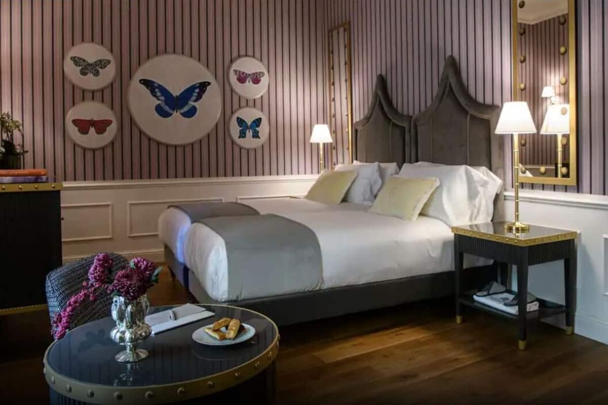 Habitación de hotel con cama, mesita de noche, lámpara y un cuadro decorativo de mariposas.