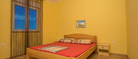 A room with a wooden bed, a red bedspread, a nightstand, a small bedside table, and a window with curtains.