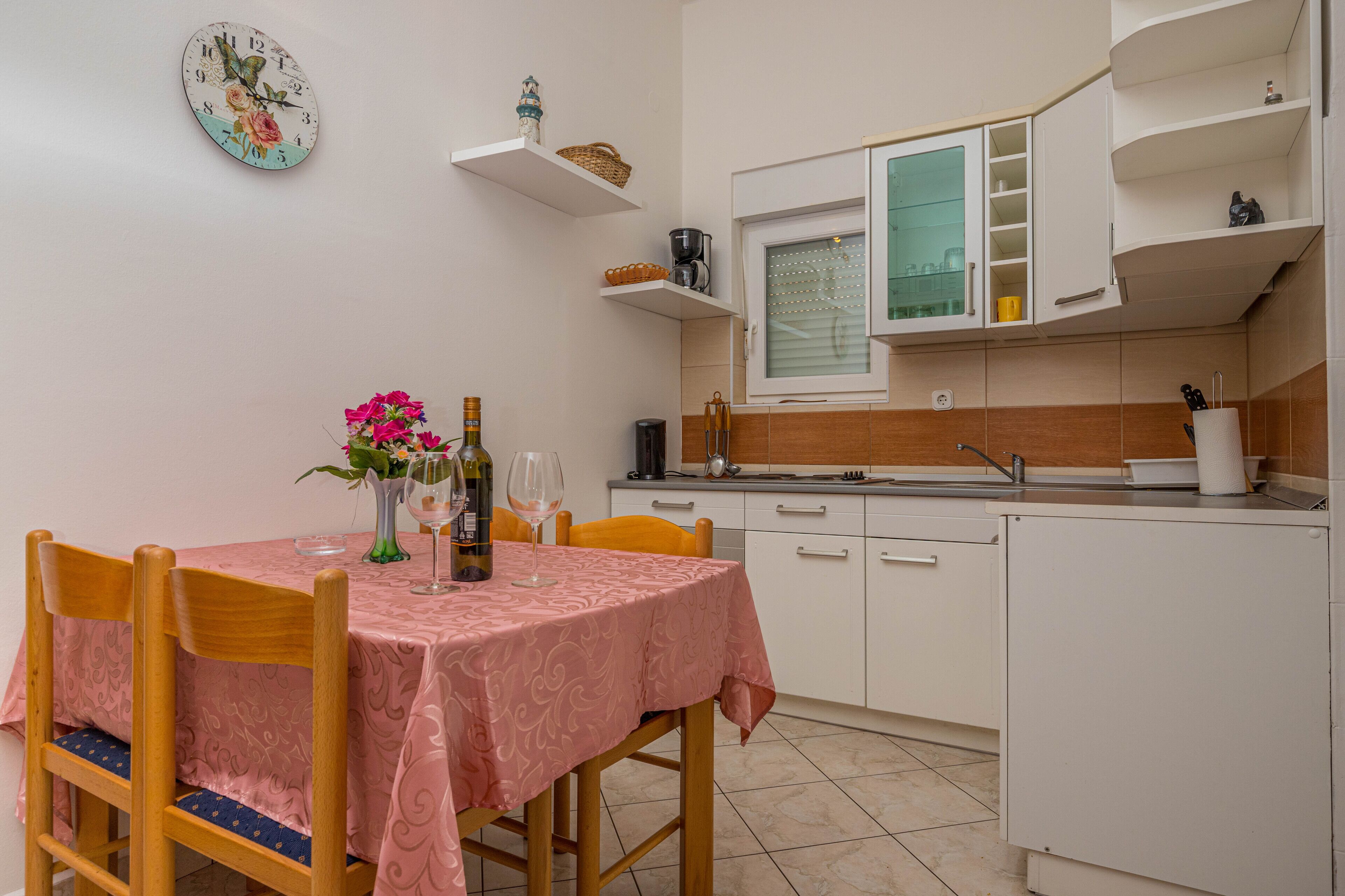 Une cuisine de taille réduite avec des meubles blancs, une table à manger recouverte d’une nappe rose et une horloge au mur.
