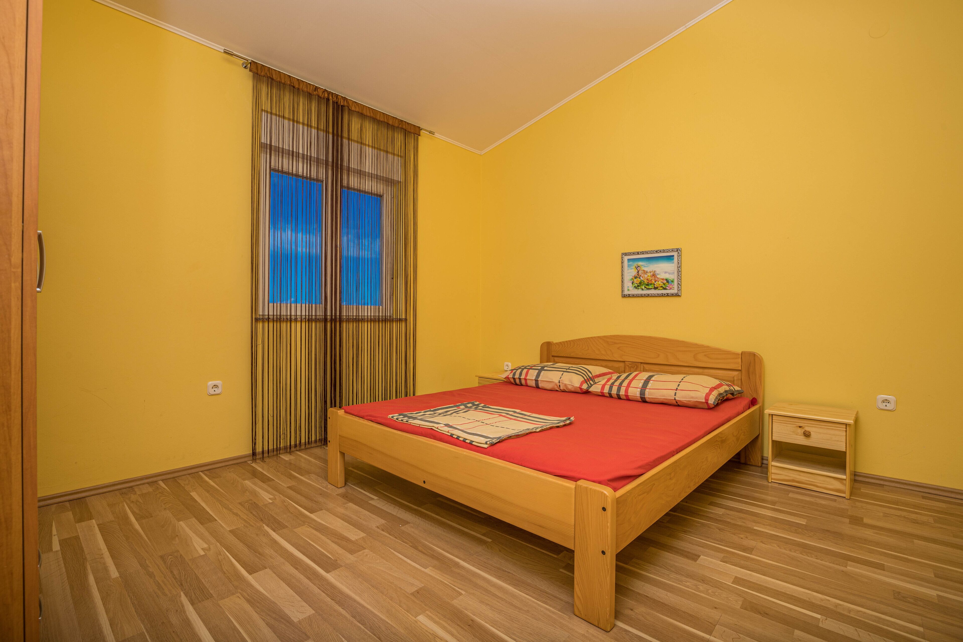 Un dormitorio con cama de madera, colcha roja y ventana con cortinas.