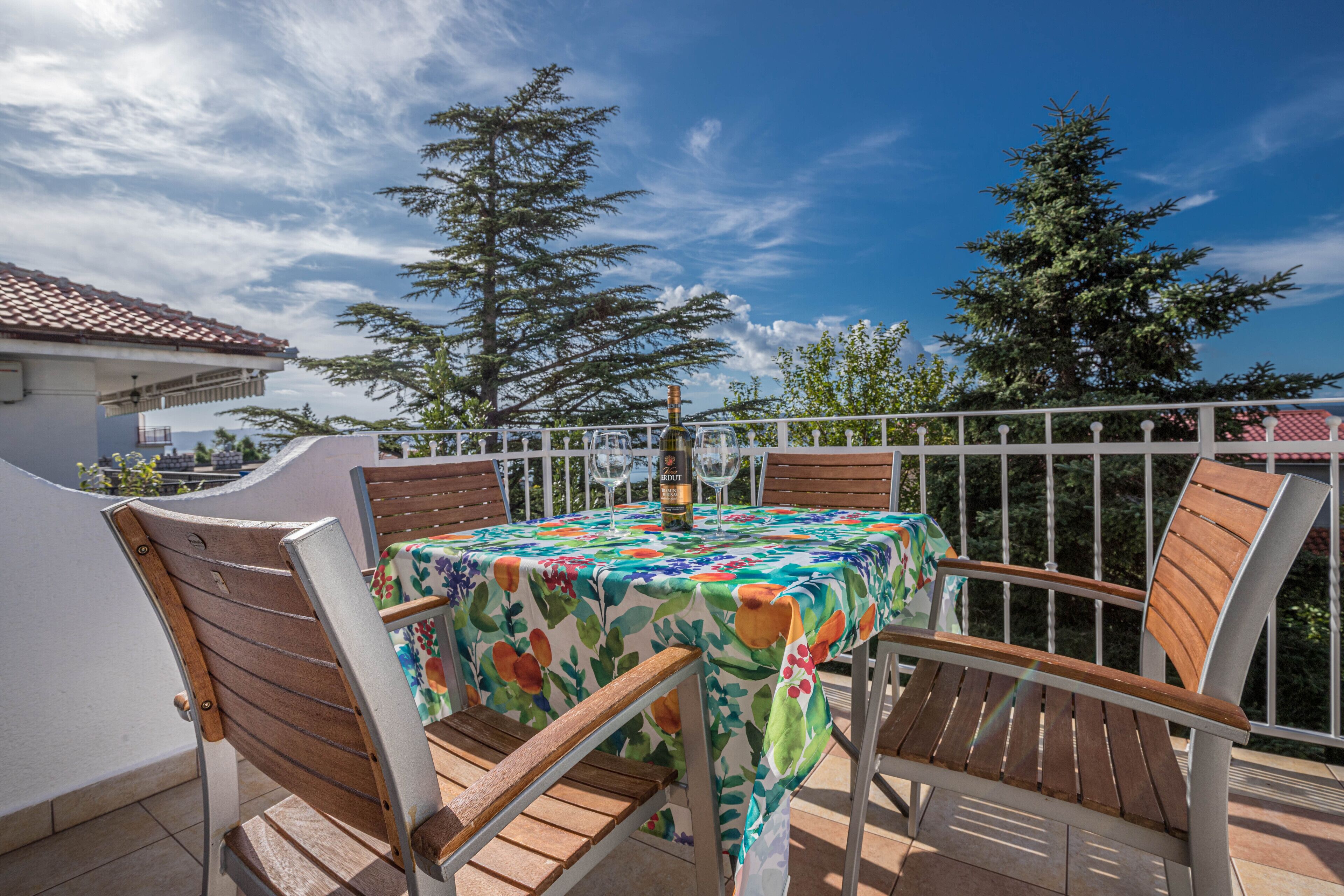A patio with a table covered in a floral tablecloth, surrounded by wooden chairs, under a clear sky.