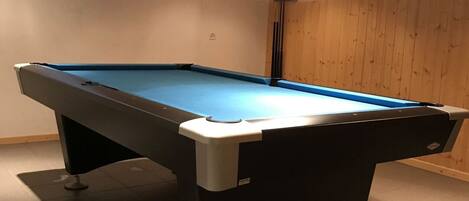 Une table de billard se trouve dans une pièce aux murs et au plafond en bois, avec trois lampes suspendues au-dessus et une petite fenêtre sur le mur.