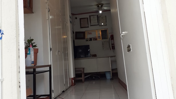 A narrow hallway with a tiled floor, a ceiling fan, and a light fixture. There is a small table with a potted plant and a bottle on it. A mat is placed on the floor near the entrance. - Posada Reforma (Oaxaca)