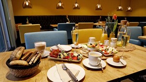 A dining table set with various dishes, including bread, fruits, and a glass of wine, in a restaurant with blue chairs and hanging lights. - Casa Palatina Vinotel (Grosskarlbach)