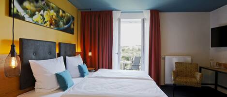 Habitación de hotel con dos camas, un ventanal grande con cortinas, una silla, una mesa con lámpara y un televisor en la pared.