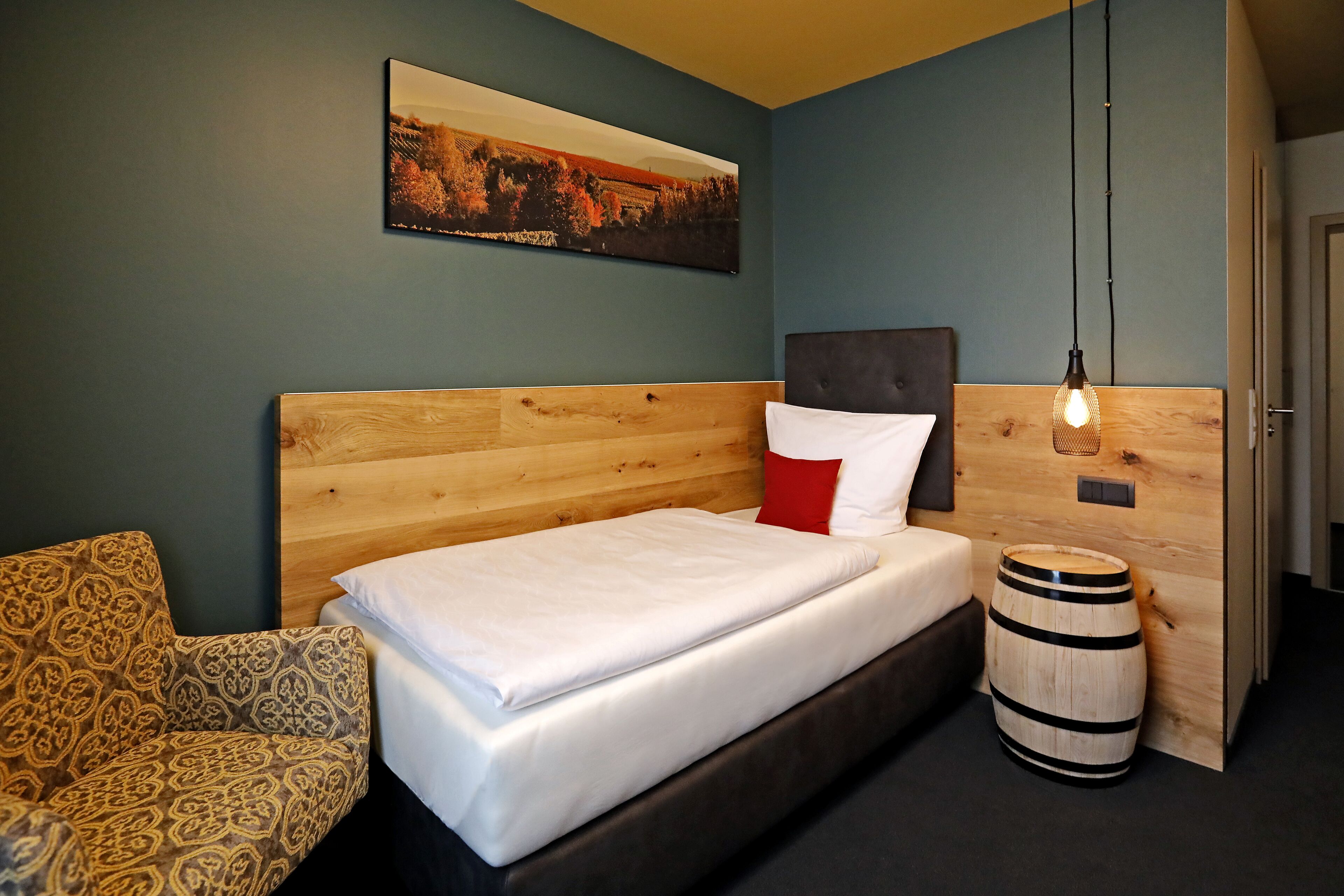 A bedroom with a wooden headboard, a bed with white bedding, a patterned armchair, a wooden barrel, and a painting on the wall.