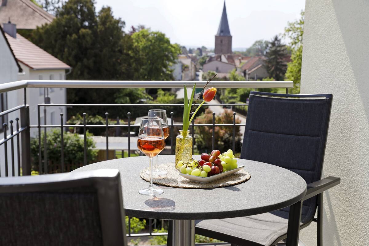 Una mesa redonda con una copa de vino, una botella y un plato de uvas en un balcón con vistas a un pueblo y una torre de iglesia.