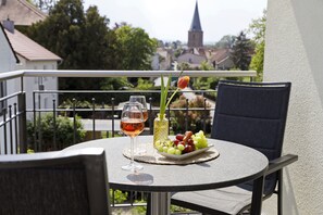 A round table with a glass of wine, a bottle, and a plate of grapes on a balcony overlooking a town with a church spire. - Casa Palatina Vinotel (Grosskarlbach)