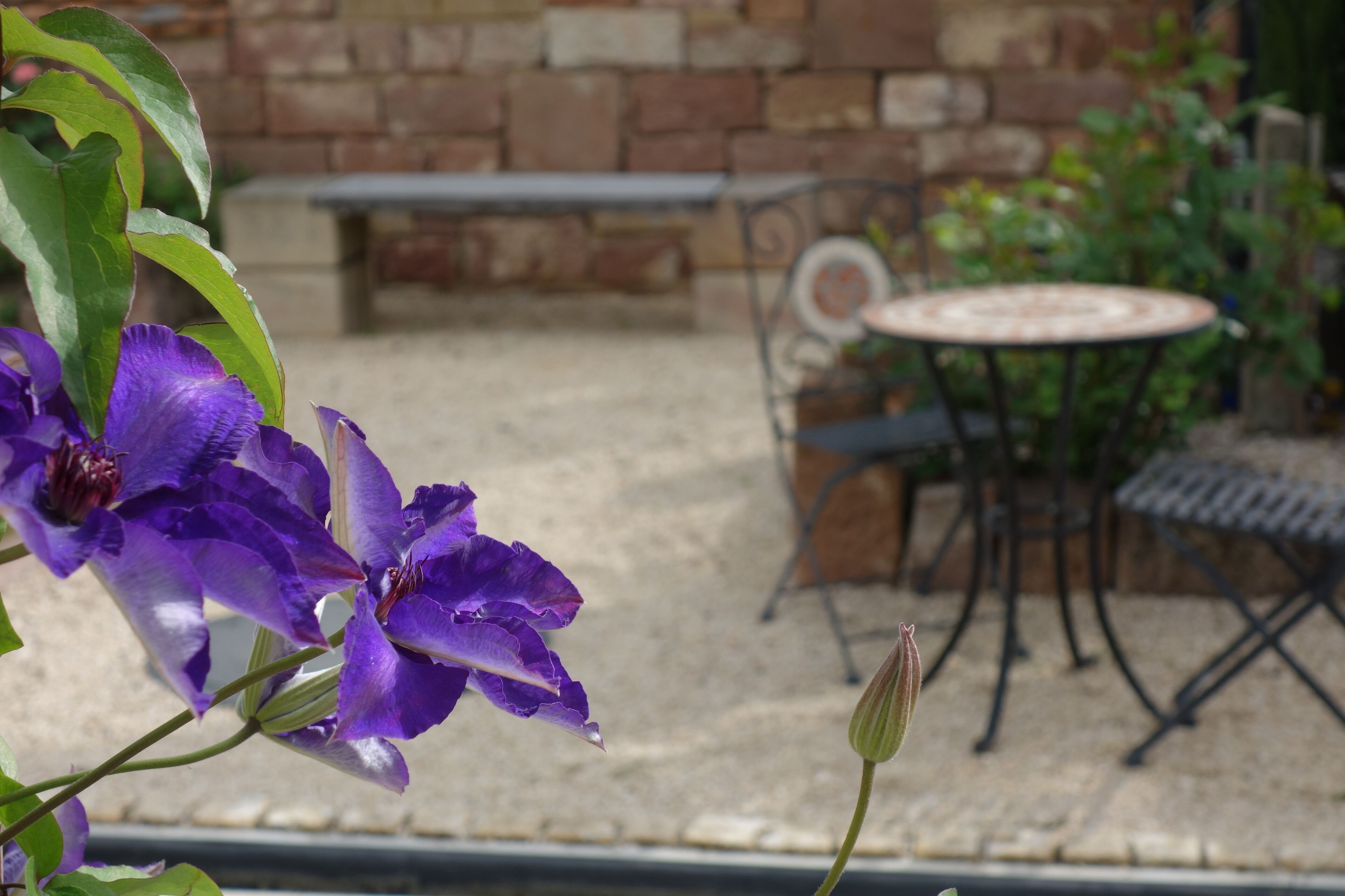 Un patio con pared de piedra, una mesa y silla de metal, y flores moradas en primer plano.