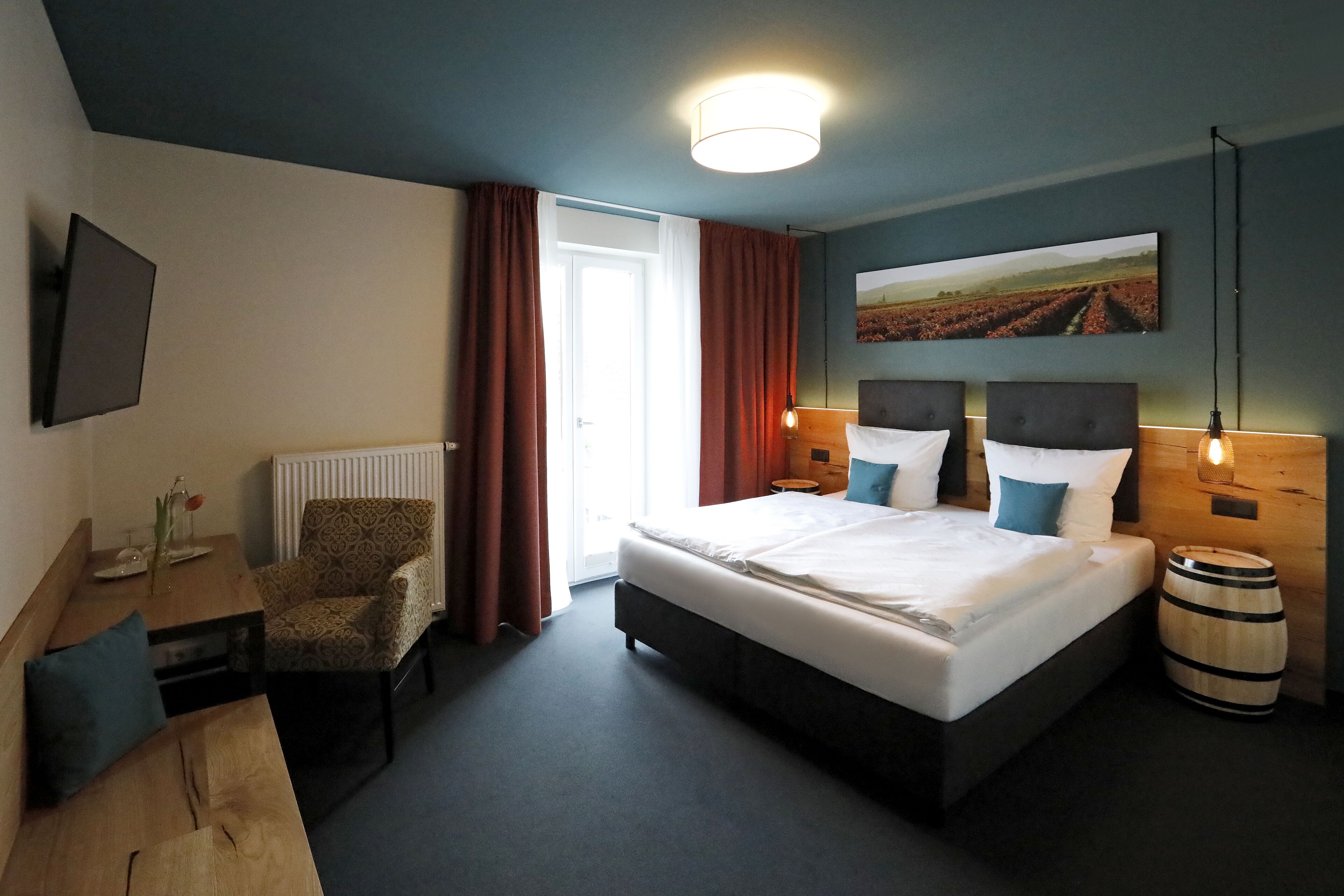 Habitación de hotel con una cama grande, un escritorio con silla, una mesita de noche de madera, un radiador, un barril de madera y un televisor montado en la pared.