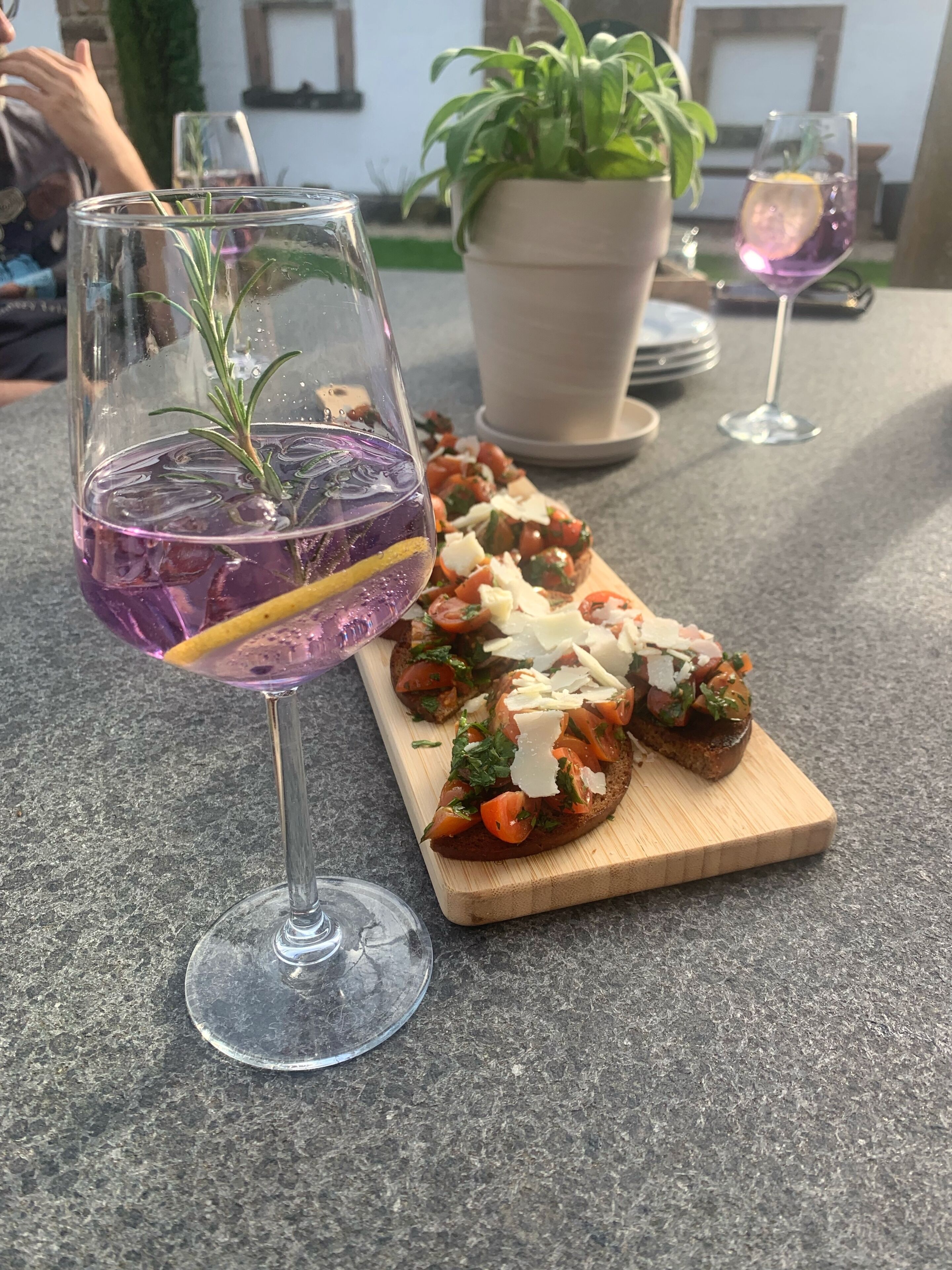 A table with two glasses of purple drink, a wooden board with food, and a potted plant in the background.