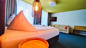 A hotel room with a bed, a sofa, a wooden barrel, a hanging light fixture, and a television. - Casa Palatina Vinotel (Grosskarlbach)