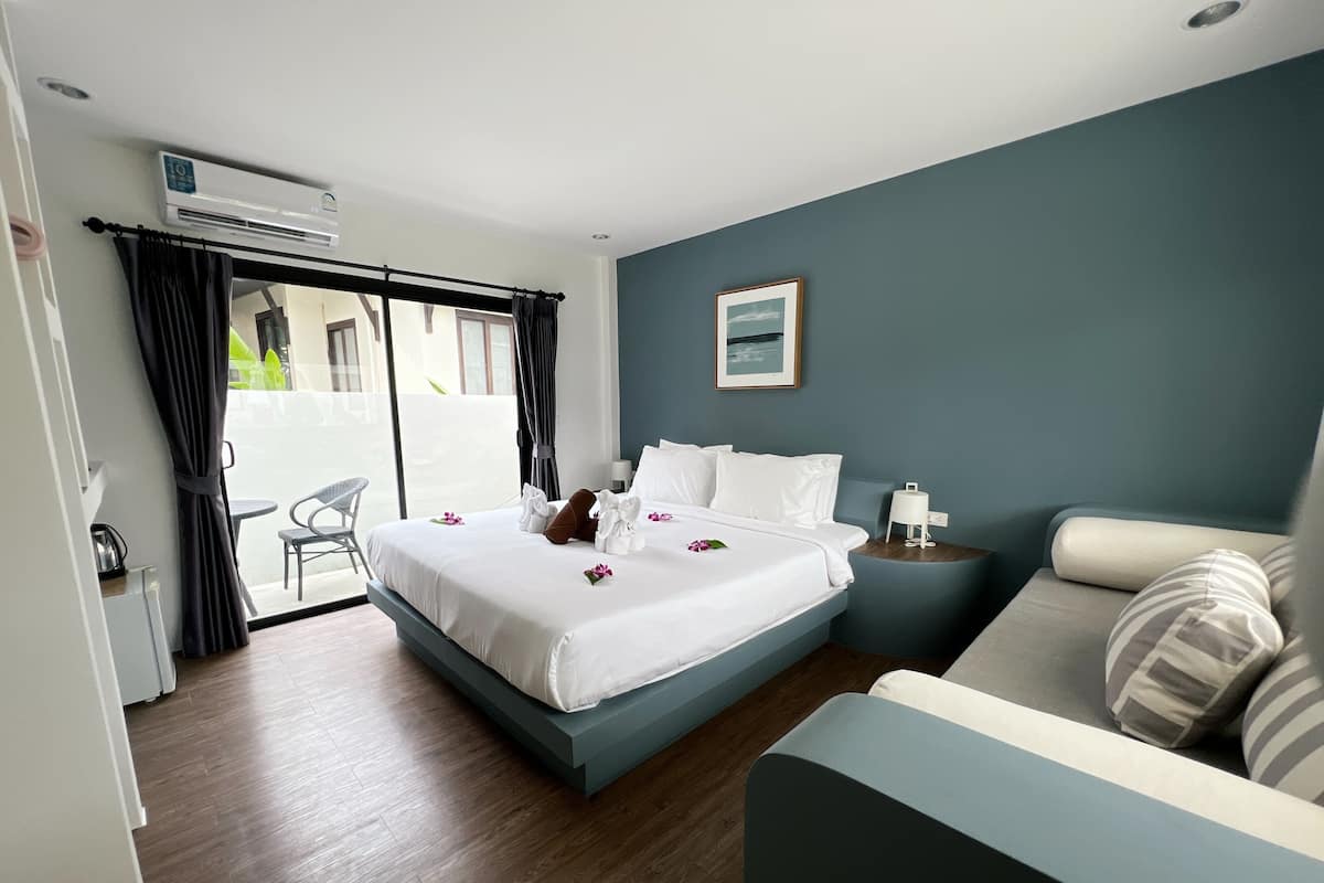 A hotel room with a bed, a bedside table, a chair, a sofa, and a view of the outdoors.