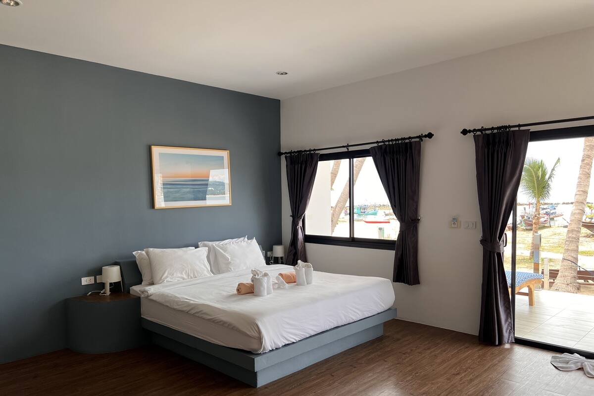 A modern hotel room with a large bed, a painting on the wall, and a view of the beach through the window.