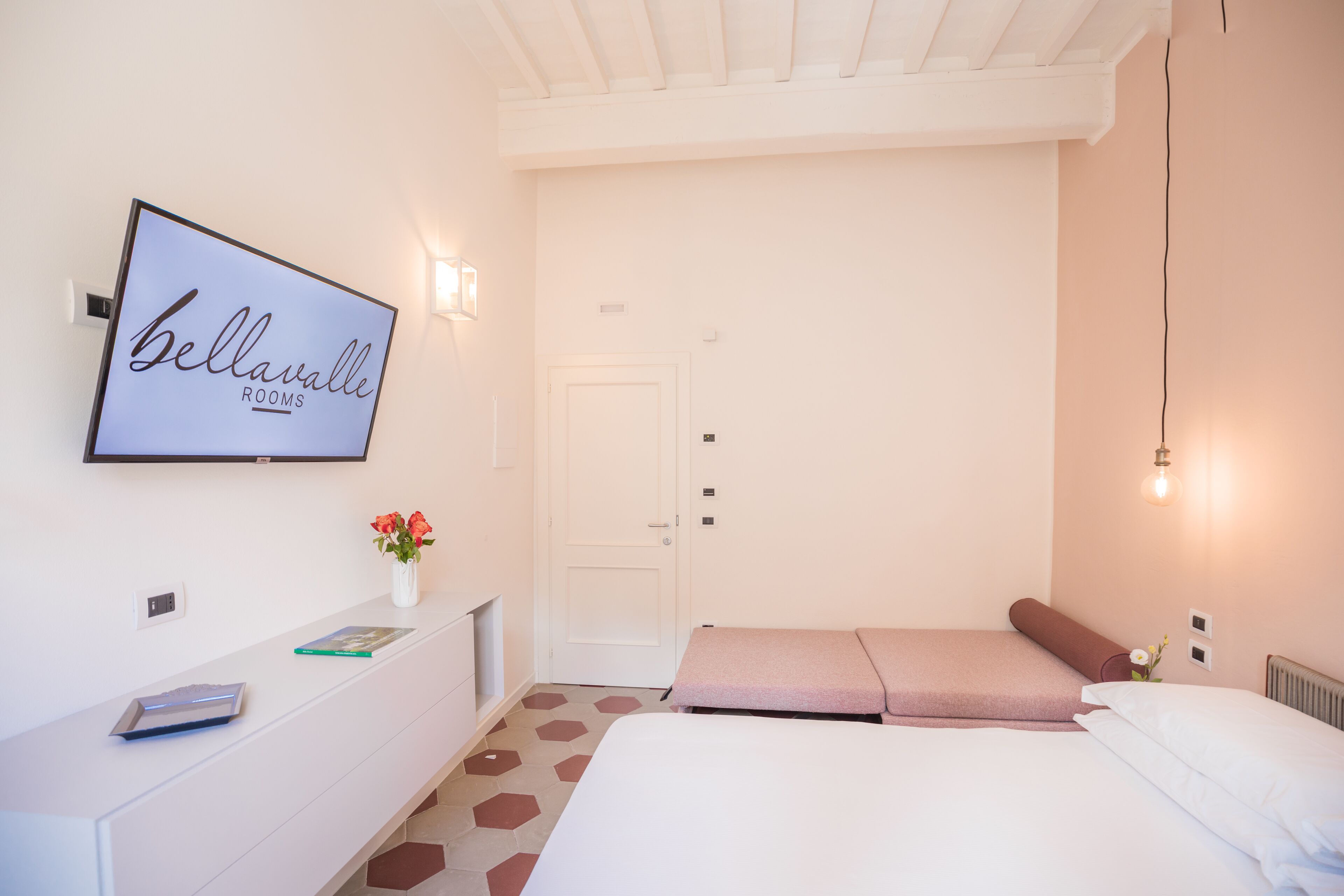 A hotel room with a bed, a bedside table with a vase of flowers, a framed sign on the wall, and a door.