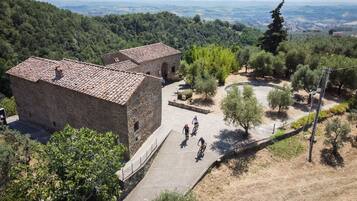 Un bâtiment en pierre avec un toit en tuiles, entouré d’arbres et bordé d’une zone pavée où se trouvent des cyclistes.