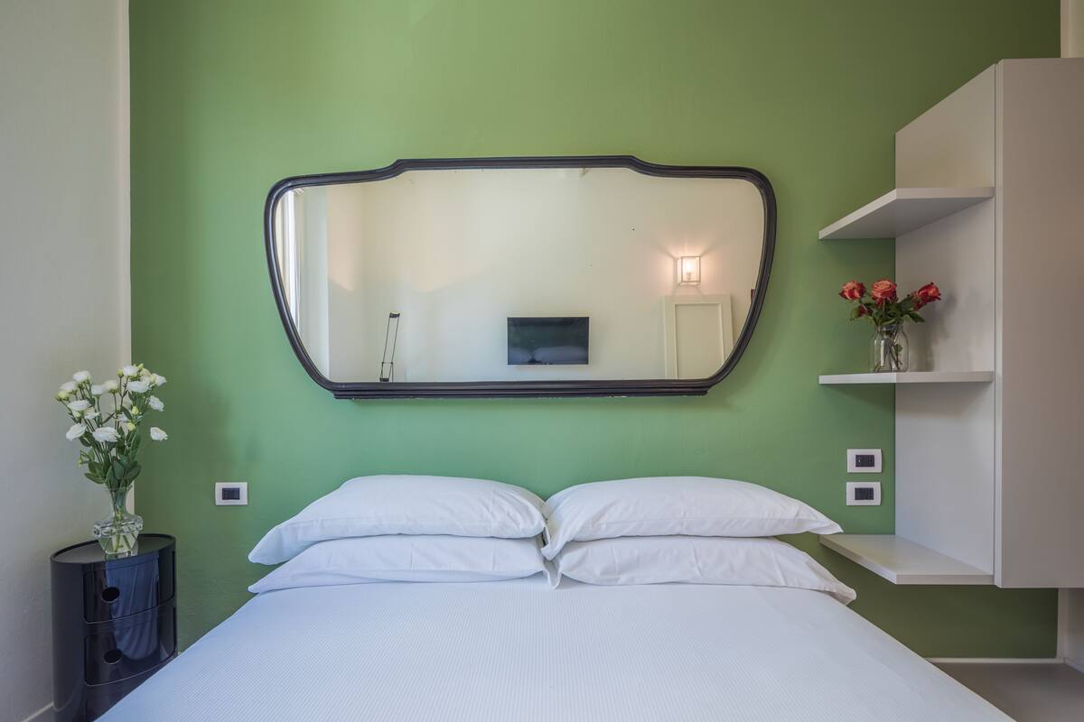 A neatly made bed with white linens, a large mirror above it, and a vase with white flowers on a bedside table.