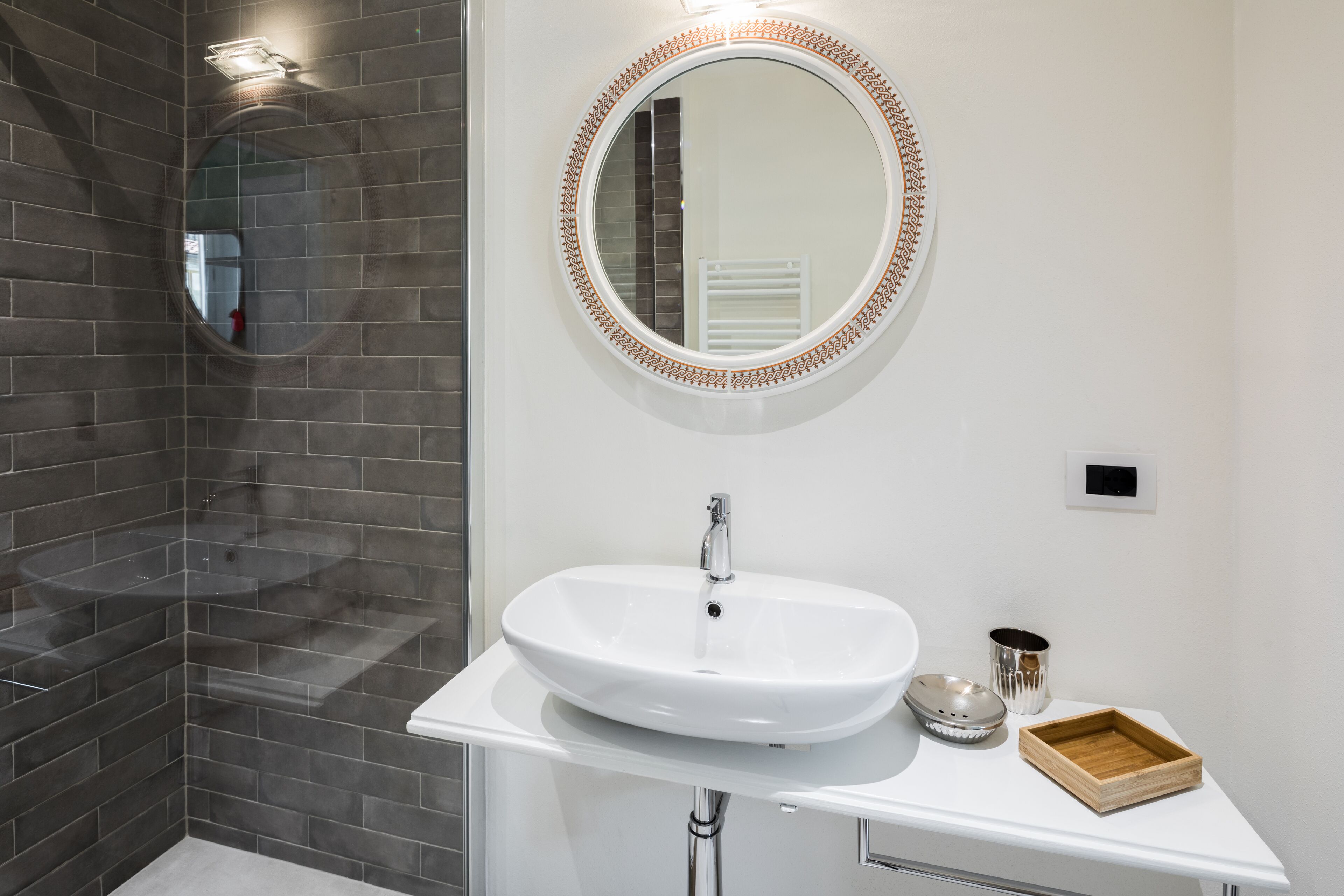 A bathroom with a white sink, a round mirror with a decorative frame, and a shower area with dark tiles.