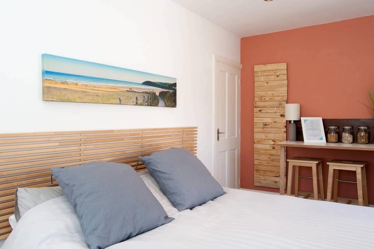 A bedroom with a bed, two pillows, a wooden headboard, a painting of a beach, a door, a wooden shelf with jars, and a small table.