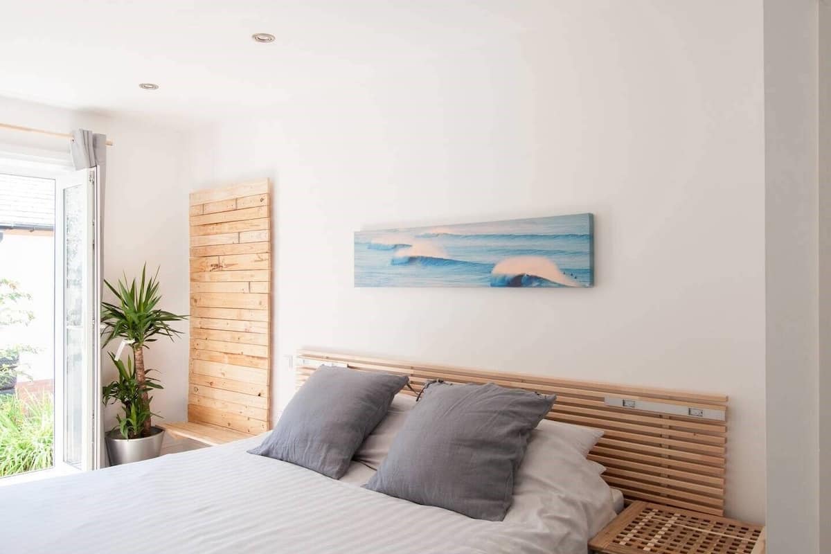 A bedroom with a wooden headboard, a bed with grey bedding, a grey pillow, a potted plant, and a painting of a wave on the wall.