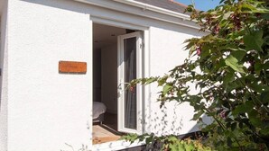 A small, white building with a door open to a room with a bed and a window. - Thistledene (Braunton)