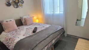 Ein Schlafzimmer mit Bett, Nachttisch, Lampe, Wandbild und einem Fenster mit Vorhängen.