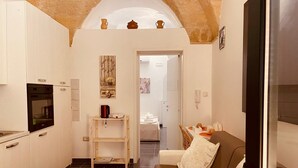 A compact kitchen with a built-in oven, a coffee machine, and a small wooden shelf. There is a brown sofa with a white cushion in the living area, and a doorway leading to a bathroom. - Suite Carlotta Premium (Lecce)