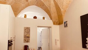 A cozy room with a stone ceiling, a wooden shelf with a kettle and a plate, a dining table with chairs, and a door leading to another room. - Suite Carlotta Premium (Lecce)