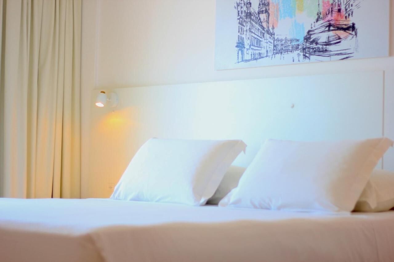 A hotel room with a bed, white curtains, a wall-mounted light, and a colorful cityscape painting.