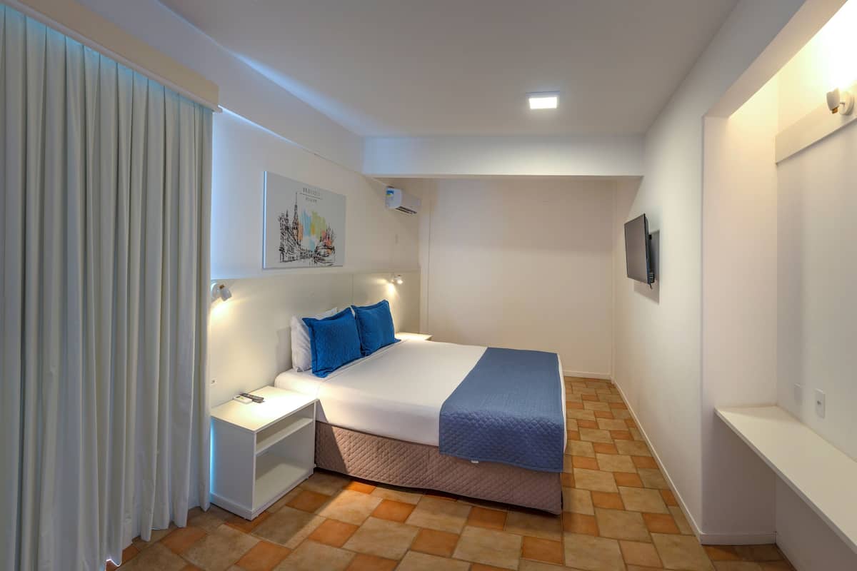 A bedroom with a bed, nightstands, a wall-mounted TV, and a window with curtains.