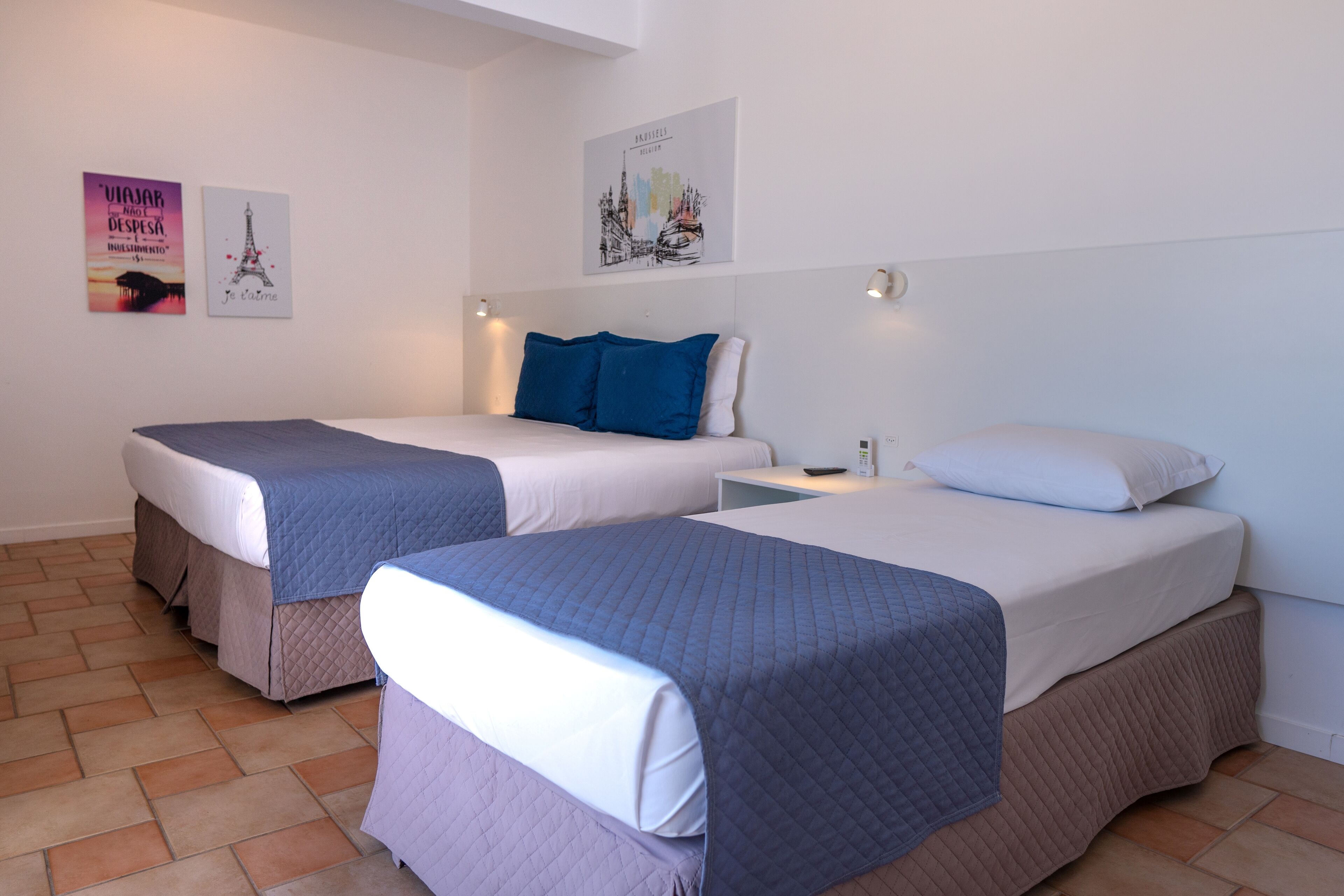 Quarto de hotel com duas camas, cada uma com colcha acolchoada e almofadas, mesa de cabeceira com abajur e obras de arte nas paredes.