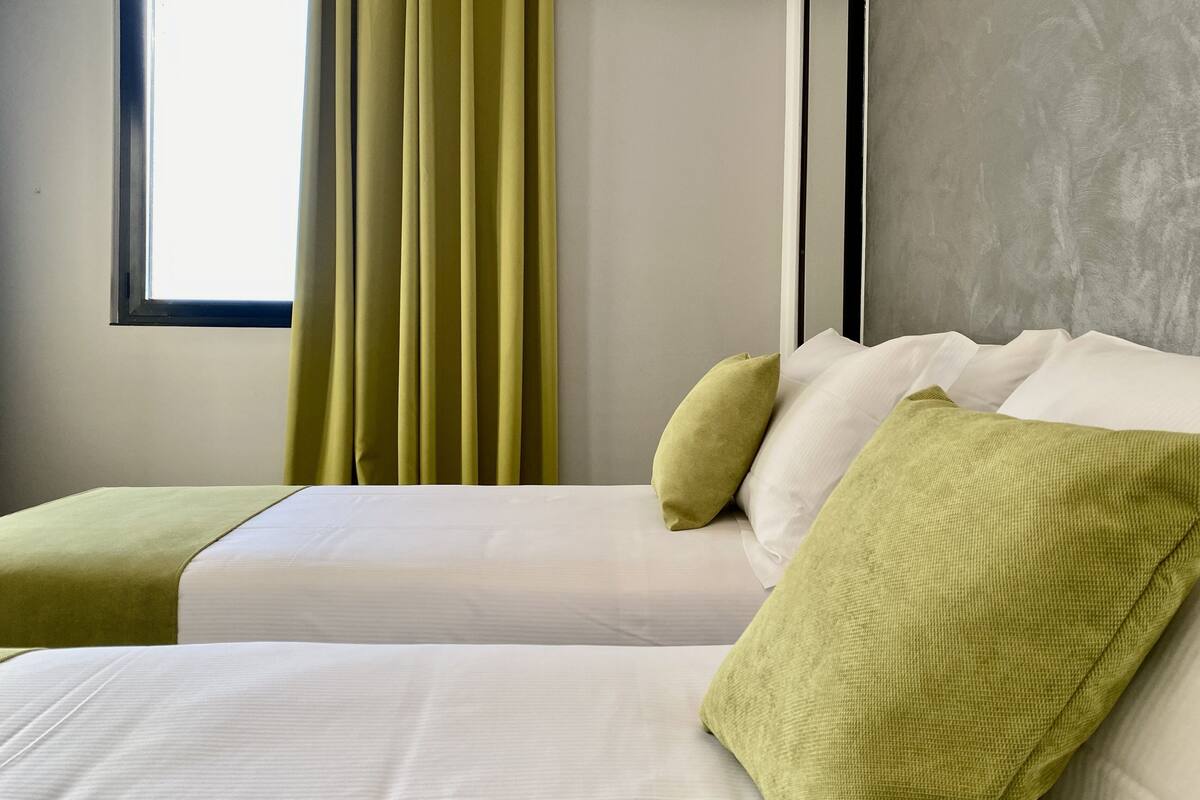 A hotel room with two beds, each with white linens and green throw pillows. There is a window with vertical blinds, and the room has a neutral color scheme with a gray accent wall.