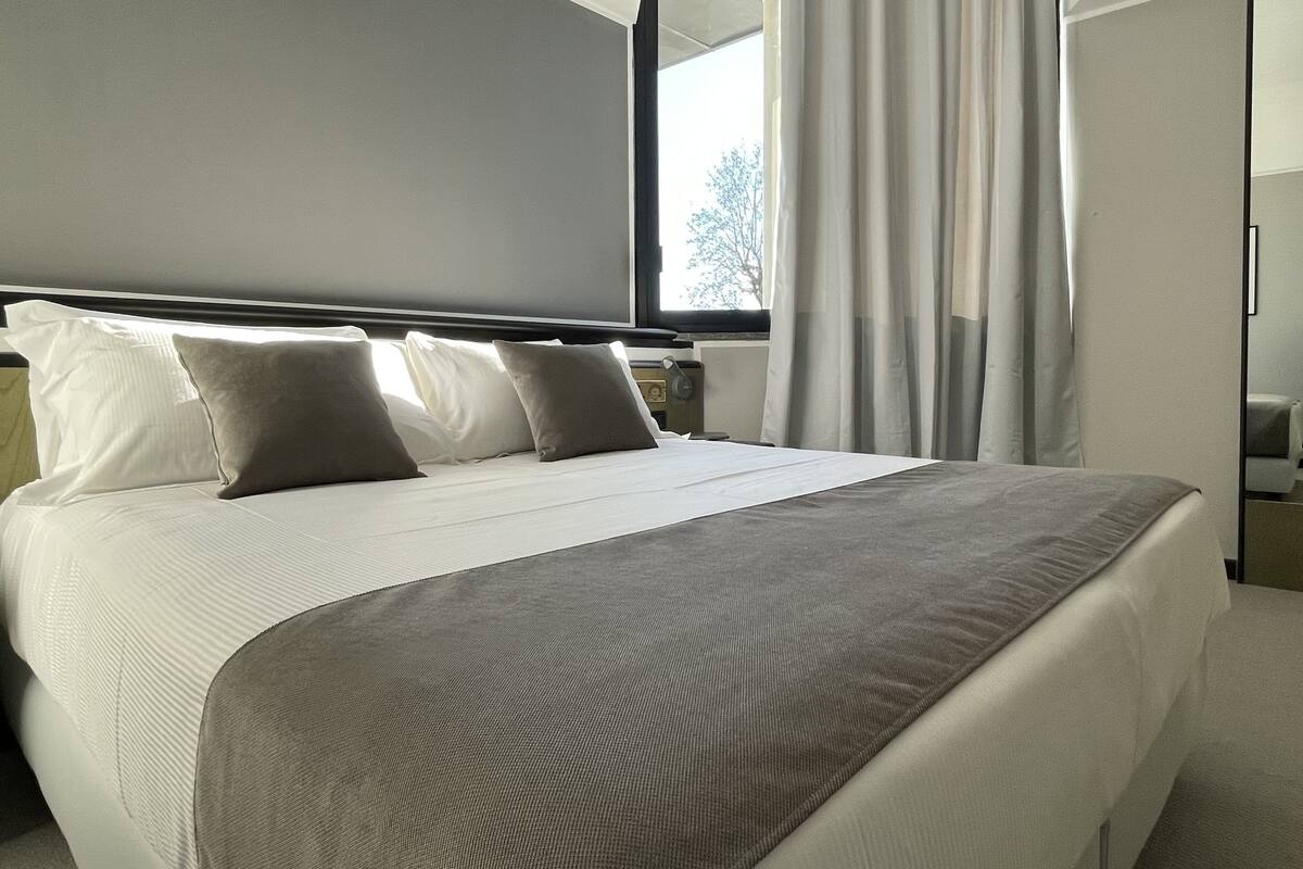 A neatly made bed with white linens and a grey blanket, a bedside table with a lamp, and a window with sheer curtains.