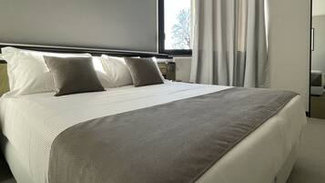 Een net opgemaakt bed met witte lakens en een grijze deken, een nachtkastje met een lamp, en een raam met dunne gordijnen.