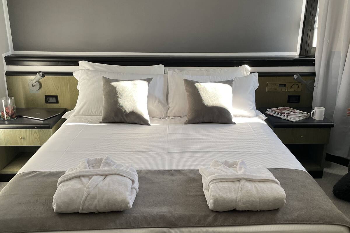 A hotel room with a large bed, two pillows, a bedside table with a lamp, a magazine, and a glass of water.