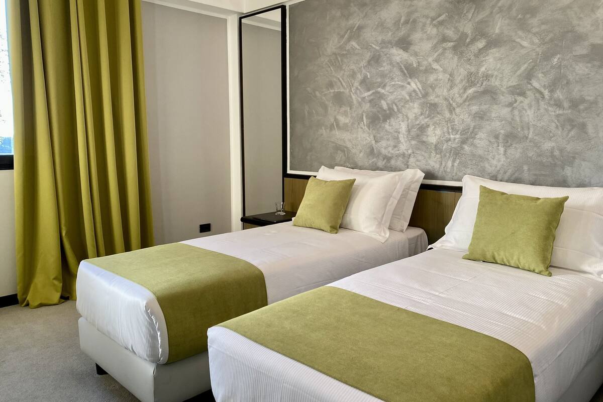 A hotel room with two beds, green and white bedding, a grey textured wall, and a large window with curtains.