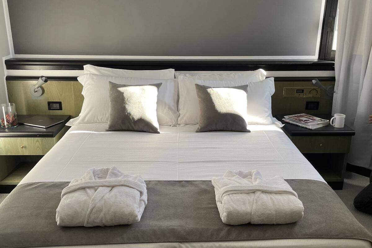 A hotel room with a bed, pillows, a bedside table with a lamp, a nightstand with a phone, and a window with curtains.