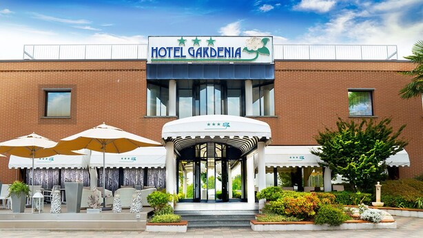 A hotel entrance with a sign reading "HOTEL GARDENIA," outdoor seating with umbrellas, and a well-maintained garden area. - Hotel Gardenia Fiera (Cermenate)