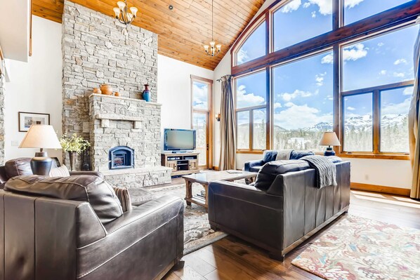 Sala de estar con chimenea de piedra, sofás de cuero, televisor y ventanas grandes con vista a las montañas.