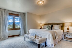 Une chambre avec une grande fenêtre, un lit avec une tête de lit, un banc, une table de chevet et une lampe.
