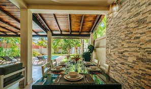 Uma sala de jantar com uma mesa de vidro posta para seis pessoas, rodeada por cadeiras brancas, sob um teto de madeira.