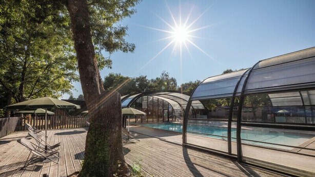 Una terraza de madera con piscina, zonas de estar cubiertas y un gran árbol.