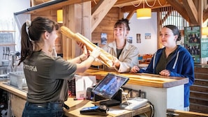 Una persona sosteniendo una baguette en un mostrador con una pantalla digital y un panel de menú al fondo.