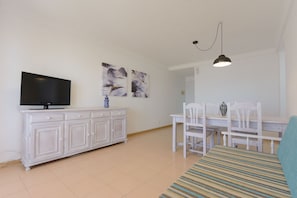 A living room with a TV on a white cabinet, a dining table with chairs, and a hanging light fixture.