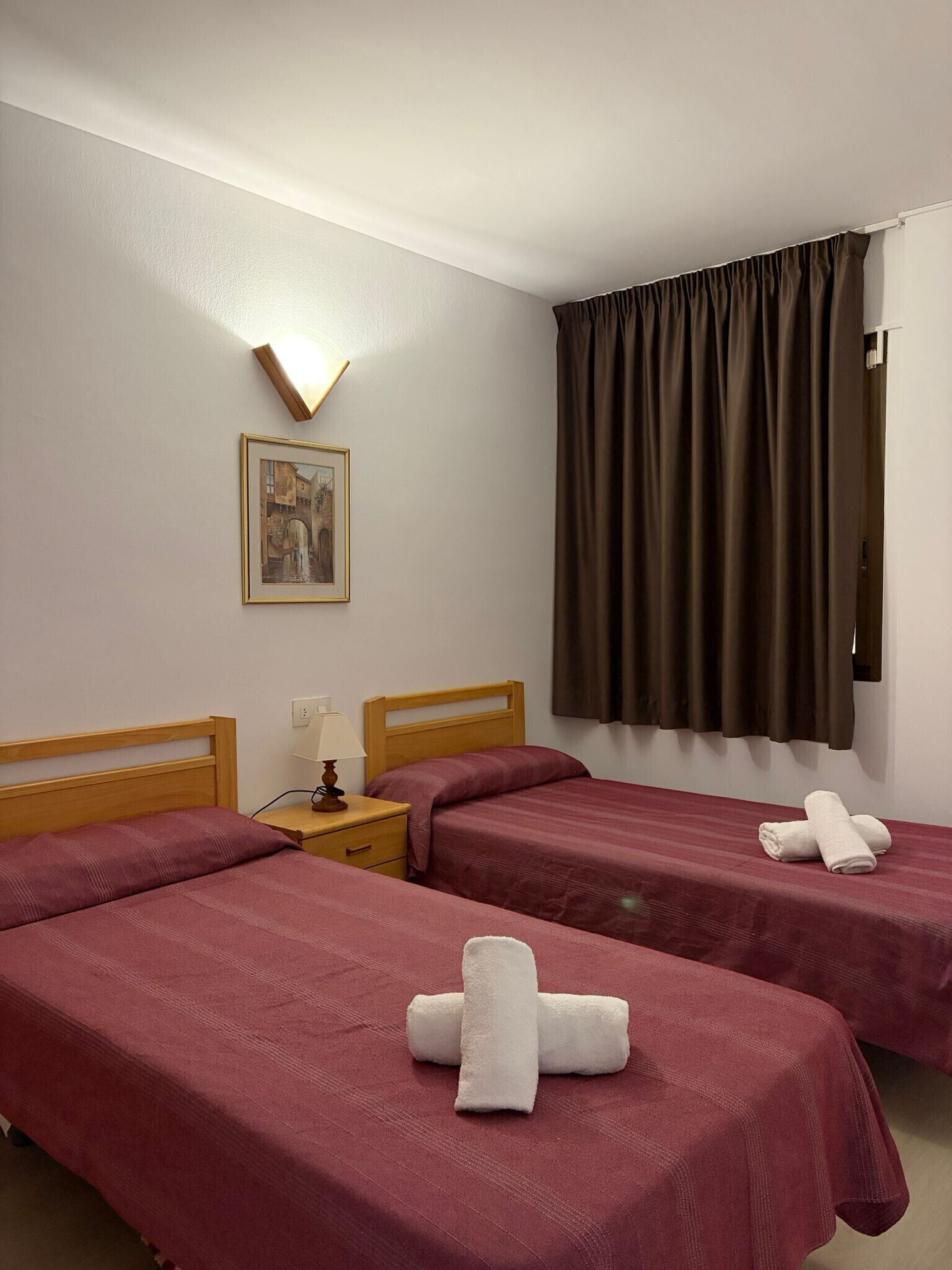 Ein Hotelzimmer mit zwei Betten, einem Nachttisch mit Lampe, einem gerahmten Bild an der Wand und einem Fenster mit Vorhängen.