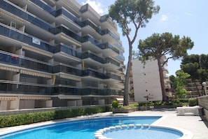 A modern multi-story building with balconies, a swimming pool, and surrounding greenery.