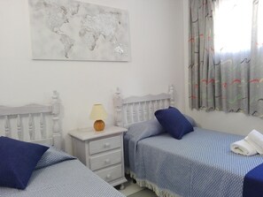 A bedroom with two beds, a nightstand, a lamp, a world map on the wall, and a window with curtains.