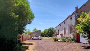 Uma entrada de cascalho leva a um edifício branco com porta vermelha, cercado por vegetação e um carro estacionado.