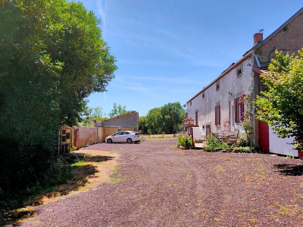 A gravel driveway leading to a white building with a red door, surrounded by greenery and a parked car. - Domaine de Cyrcé (La Celle)