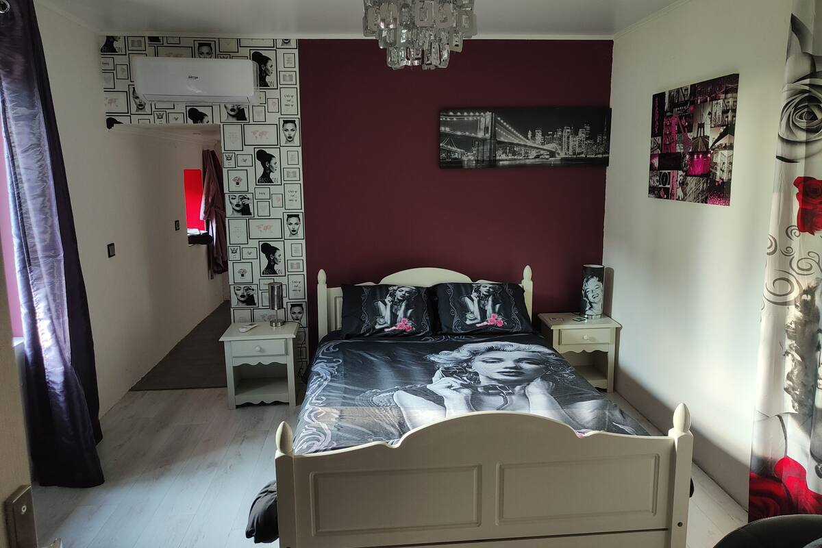 Un dormitorio con cama, mesitas de noche, una lámpara de araña, una pared con cuadros enmarcados y una pared de acento en rojo.